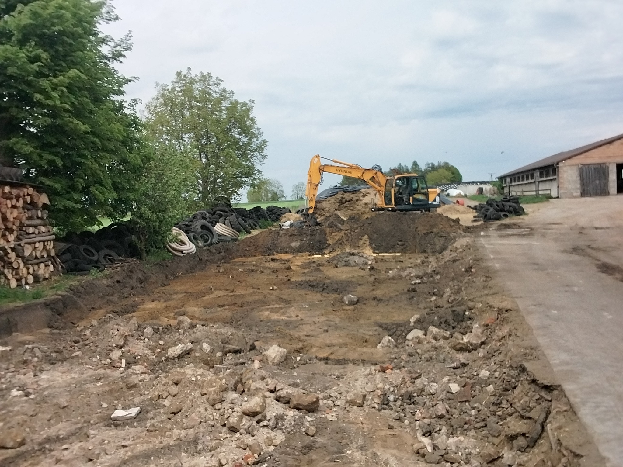 Żółta koparka Hyundai podczas prac ziemnych na placu budowy. W tle sterta opon i budynek gospodarczy. Widoczne ślady świeżo przekopanej ziemi i gruz.