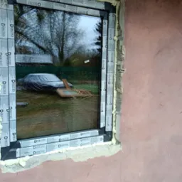 Montaż okna Drutex z widoczną pianką montażową i folią ochronną na profilach, odbicie ogrodu z przykrytym samochodem w szybie.