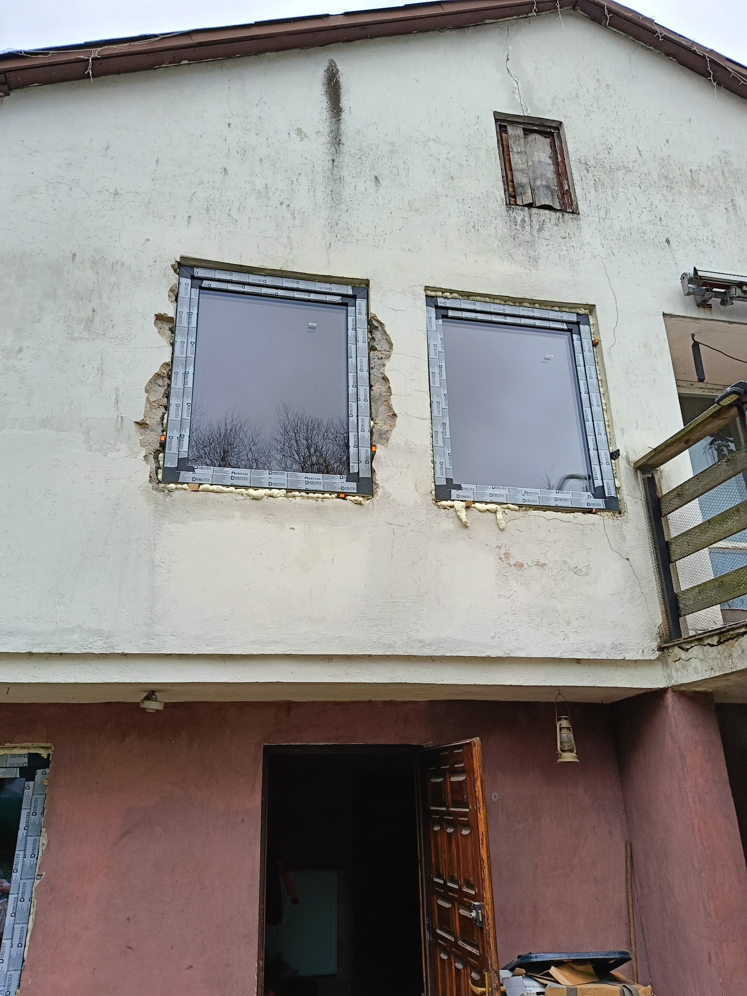 Montaż okien PCV w surowej elewacji, widoczne pianowanie i uszczelnianie, fragment starego okna zabitego deskami, budynek w trakcie remontu.