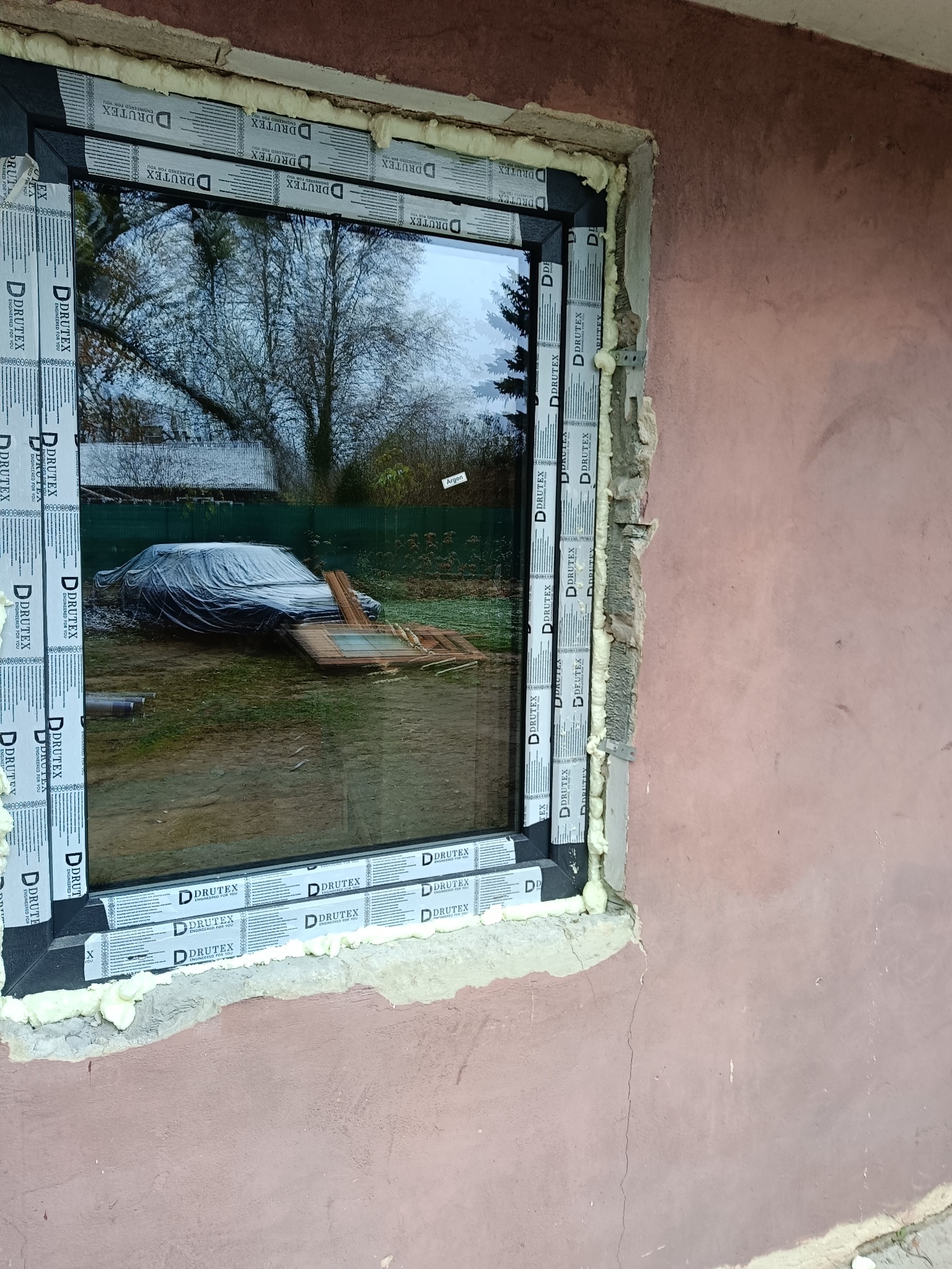 Montaż okna Drutex z widoczną pianką montażową i folią ochronną na profilach, odbicie ogrodu z przykrytym samochodem w szybie.