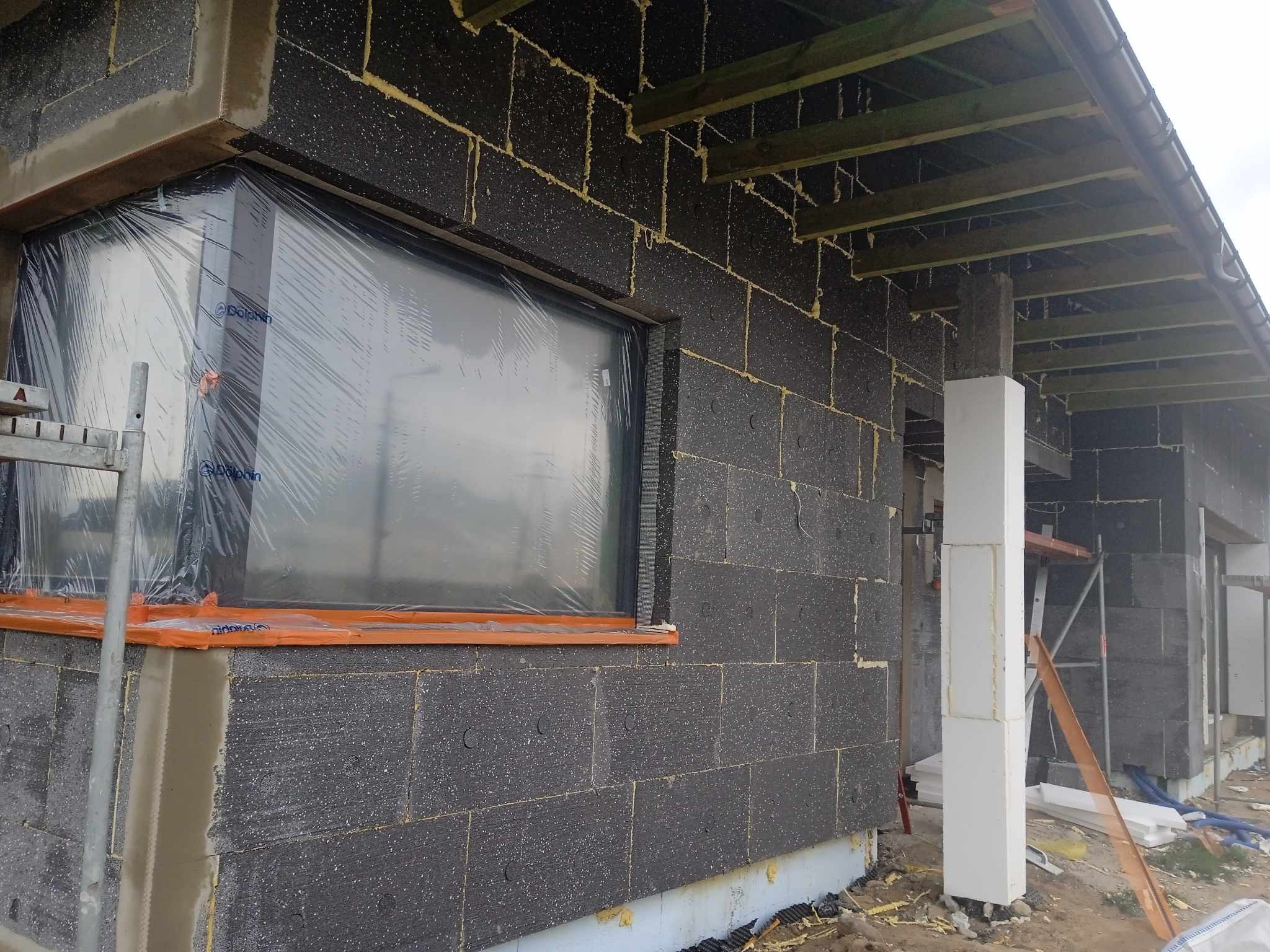 Budynek w trakcie ocieplania zewnętrznego, widoczne czarne płyty izolacyjne z pianką montażową w szczelinach, okno zabezpieczone folią, drewniana konstrukcja dachu.