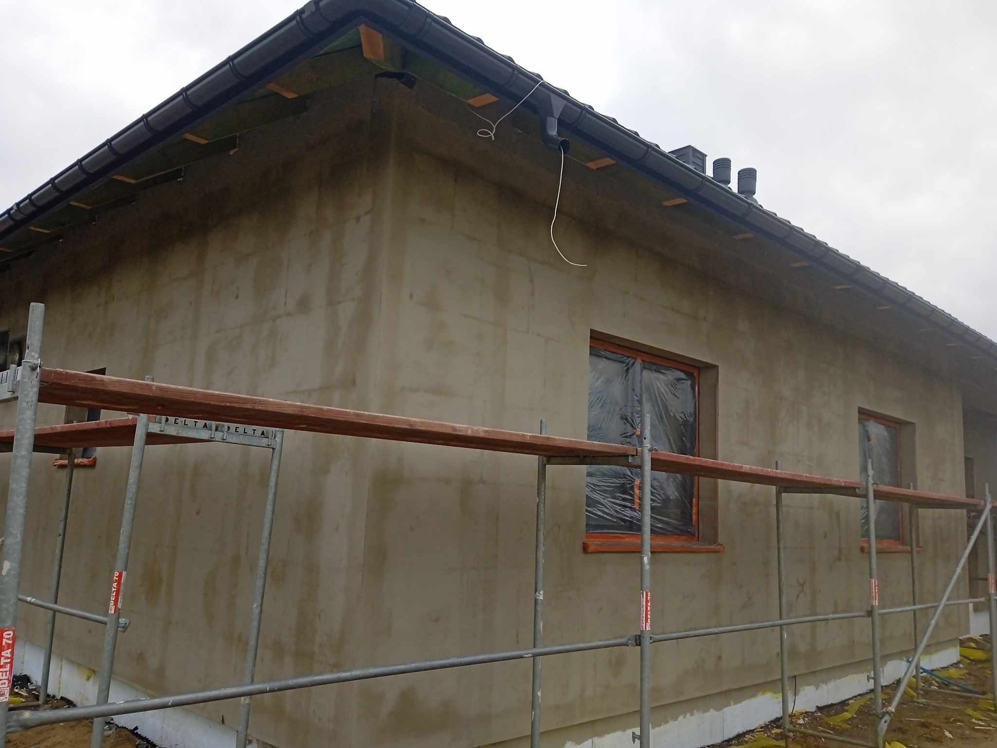 Surowa elewacja domu jednorodzinnego w trakcie prac wykończeniowych, widoczne rusztowanie, okna zabezpieczone folią, świeżo nałożony tynk w neutralnym kolorze, czarny dach z orynnowaniem.