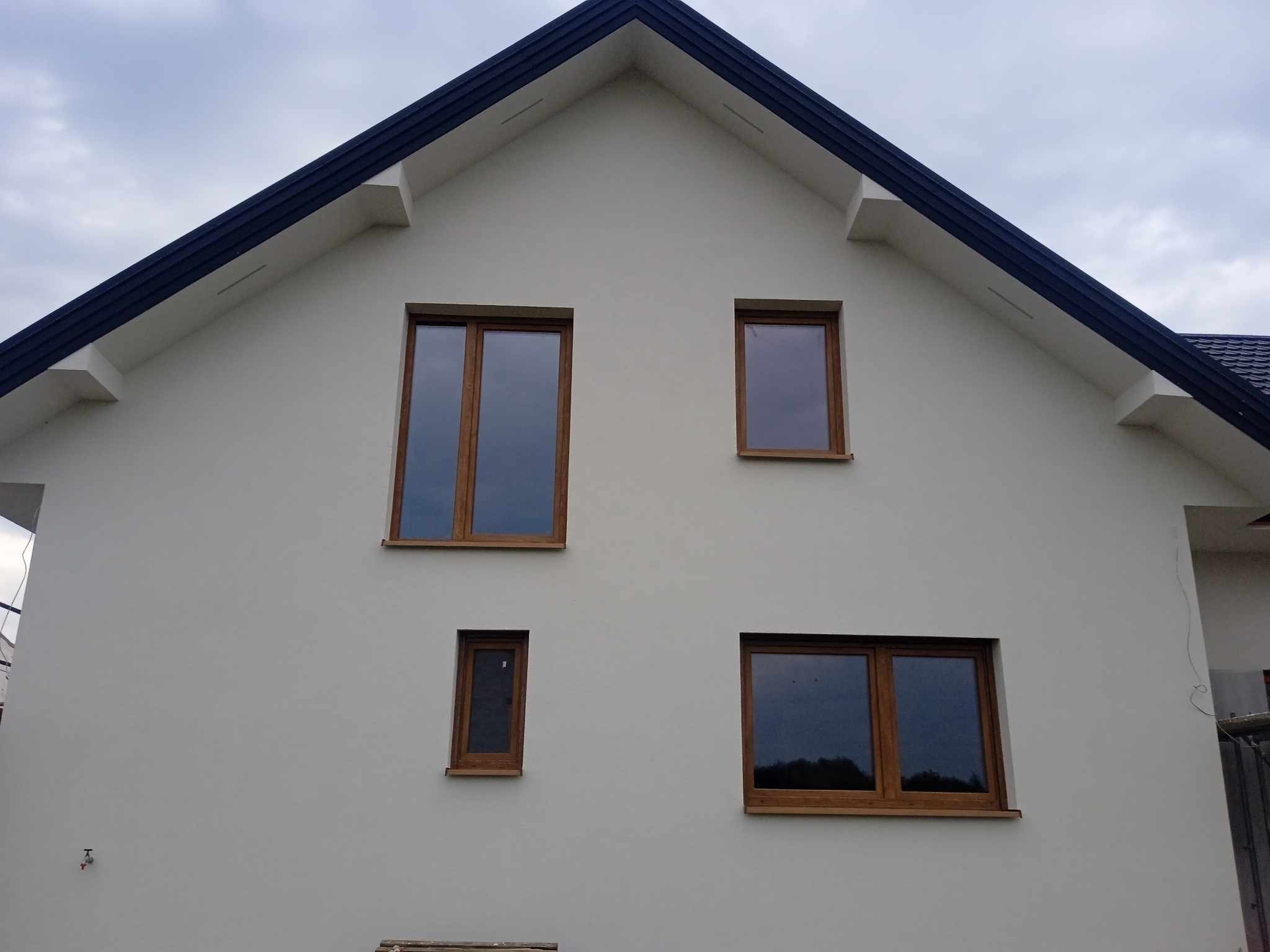 Elewacja domu jednorodzinnego po remoncie, z nowymi oknami o drewnianych ramach i świeżo pomalowaną fasadą w odcieniach szarości, widoczne gniazdko elektryczne na ścianie.