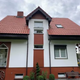 Dwukondygnacyjny dom z czerwoną dachówką, białymi ścianami z ceglanymi akcentami, dekoracjami ogrodowymi i spiralnie przyciętym krzewem.