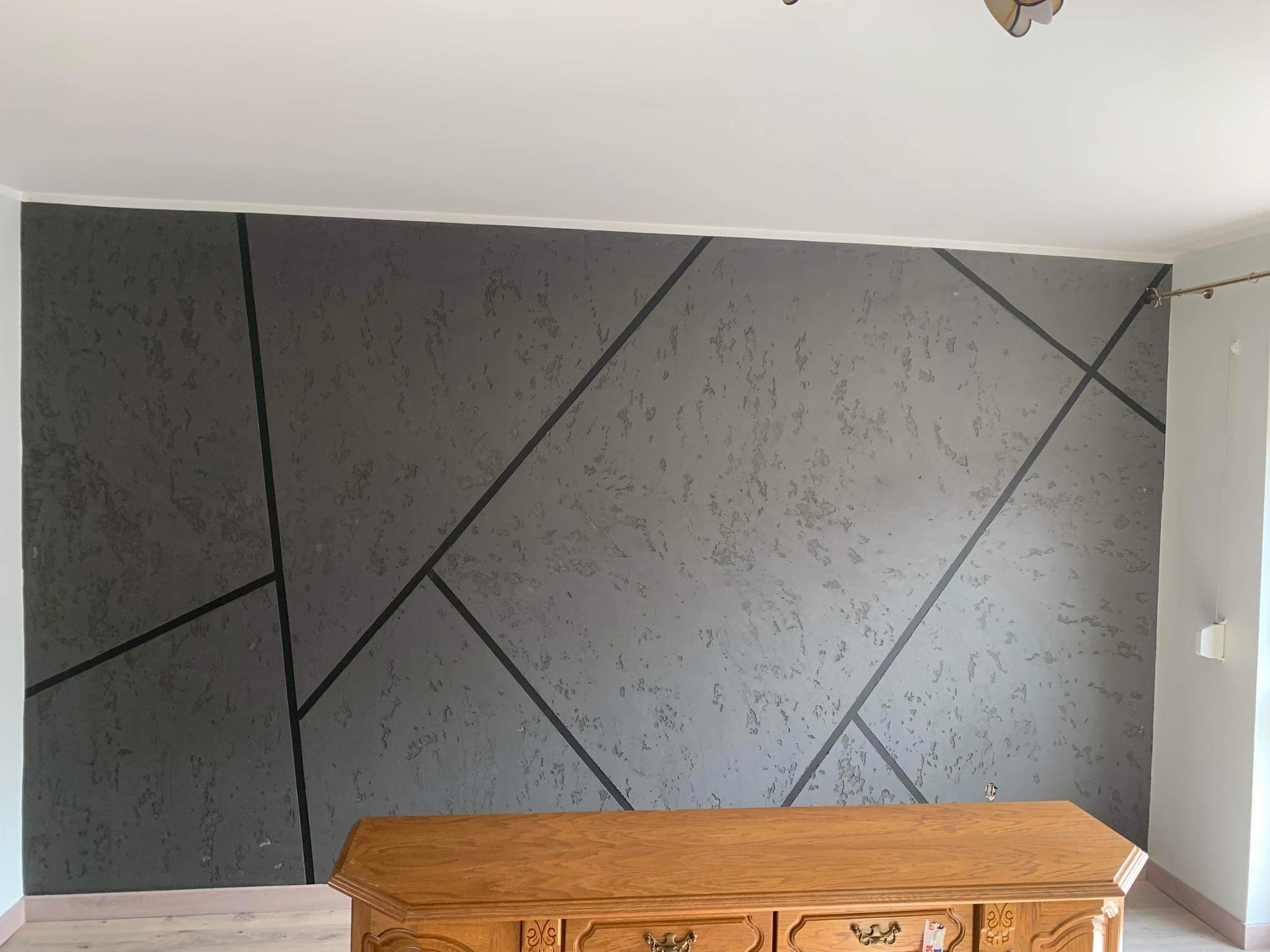 Szara ściana z geometrycznym wzorem wykonanym z ciemnych linii, poniżej widoczny fragment drewnianej komody.