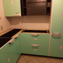 Krzysztof - Montaż jasnych, miętowych szafek kuchennych z czarnymi blatami i srebrnymi uchwytami, częściowo jeszcze zabezpieczonych folią ochronną, widoczna brązowa podłoga z płytek.