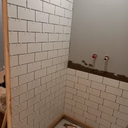 Krzysztof - Fragment łazienki w trakcie układania białych, prostokątnych płytek z użyciem krzyżyków dystansowych; widoczne wystające rury instalacyjne i narzędzia na podłodze.