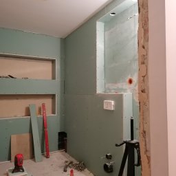 Krzysztof - Łazienka w trakcie remontu: ściany obłożone płytami gipsowo-kartonowymi, widoczne otwory na półki i instalacje, narzędzia budowlane na podłodze.