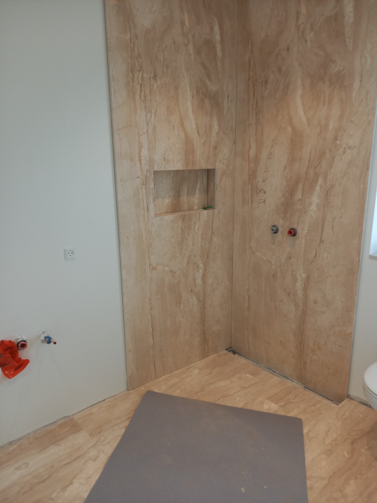 Fragment łazienki w trakcie prac wykończeniowych: ściany wyłożone płytami imitującymi trawertyn, wnęka w ścianie, przygotowane instalacje wodne, zabezpieczona podłoga.