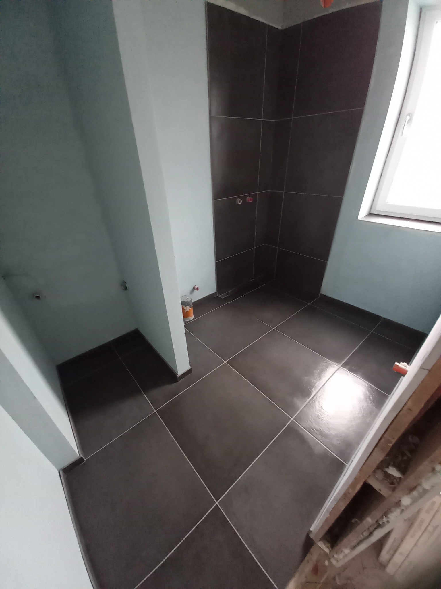 Łazienka w trakcie wykańczania, widoczne ciemne płytki podłogowe i ścienne, ściany pomalowane na jasny kolor, okno oraz fragment drewnianej ramy drzwi.