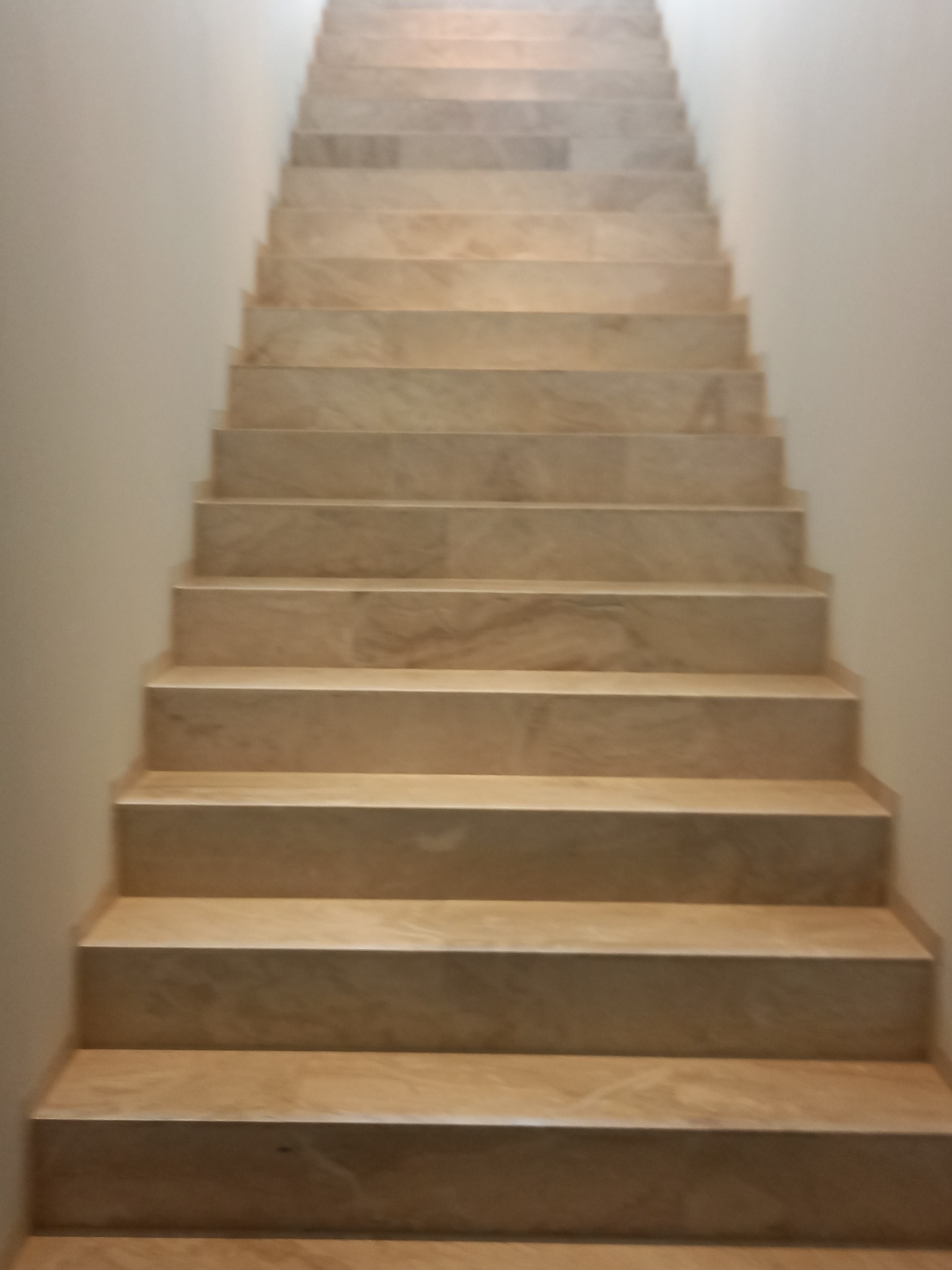 Wykończone schody wewnętrzne z beżowych płytek imitujących marmur, oświetlone od góry, widok z dołu.