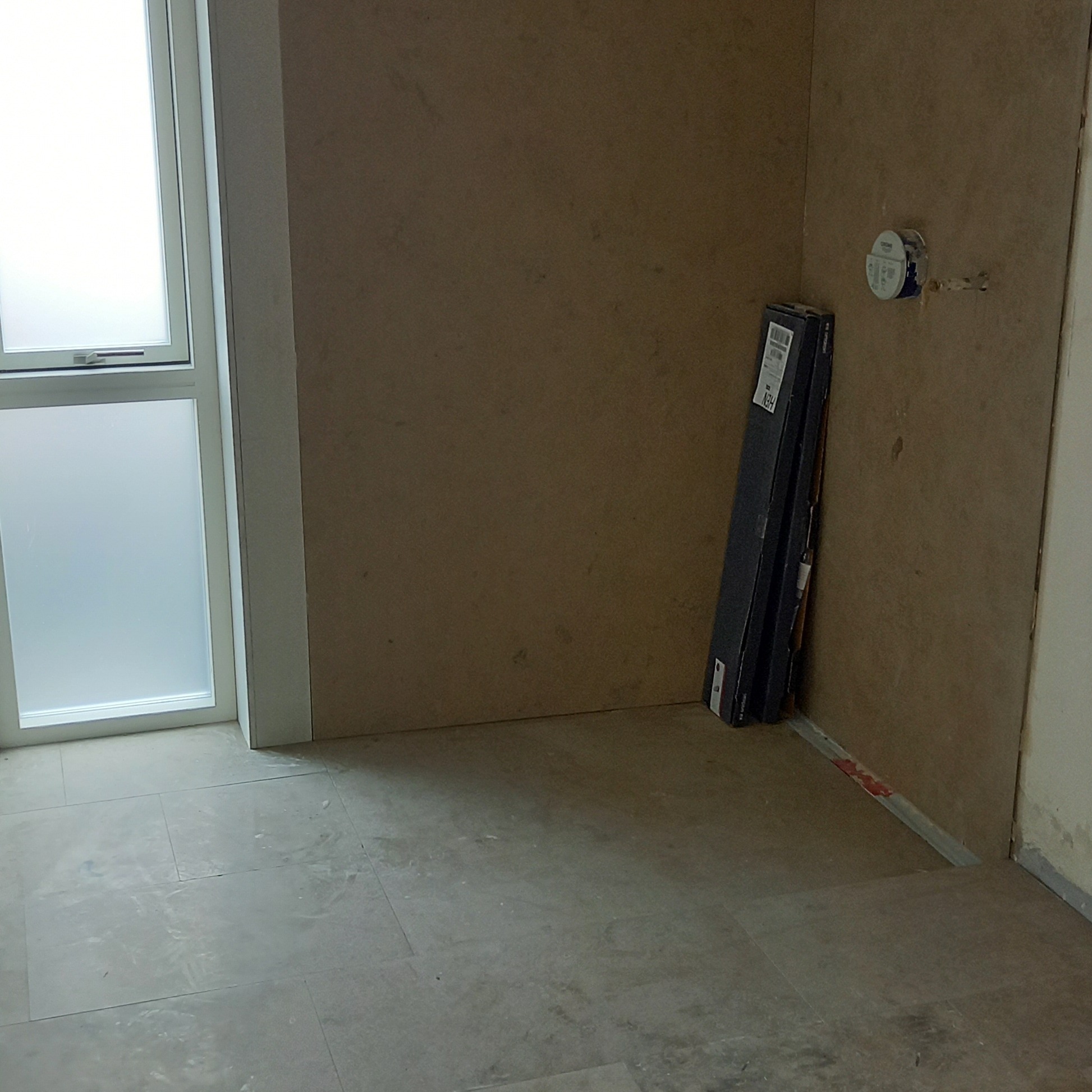 Łazienka w trakcie remontu: surowe ściany przygotowane do wykończenia, widoczne puszki elektryczne, okno z matową szybą i paczki paneli podłogowych oparte o ścianę.