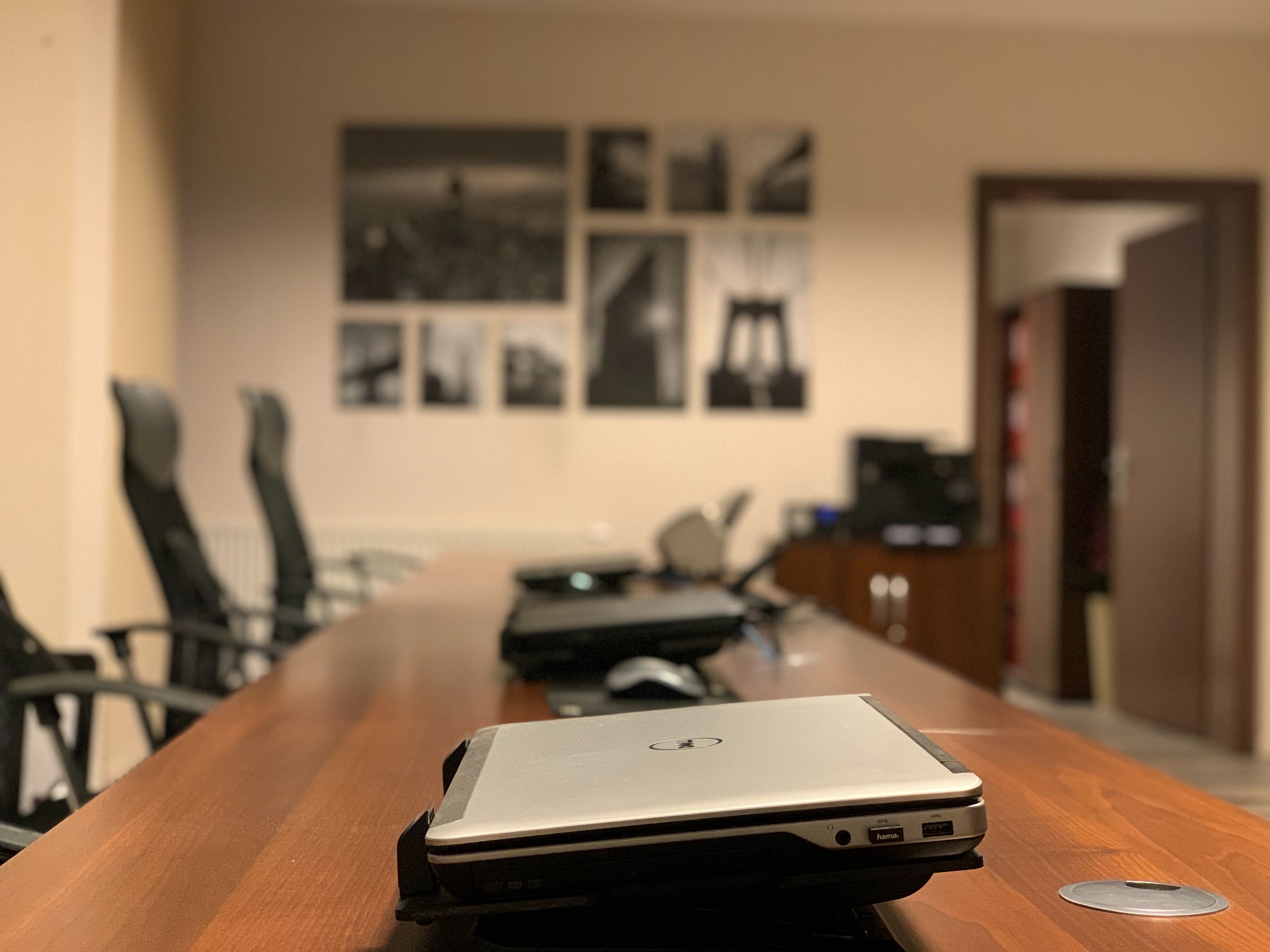 Biuro z długim drewnianym stołem konferencyjnym, na którym leżą laptopy, w tle ściana z czarno-białymi zdjęciami i otwarte drzwi do gabinetu.