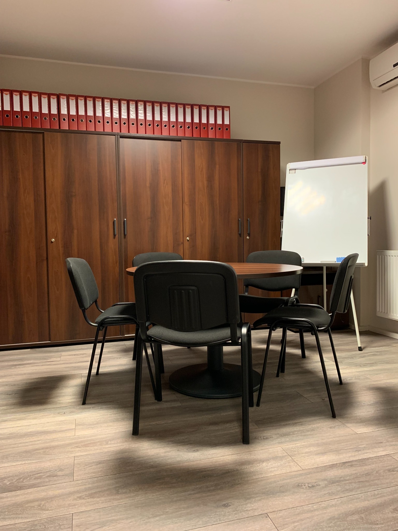 Sala konferencyjna z okrągłym stołem, czarnymi krzesłami, szafą z segregatorami i tablicą flipchart