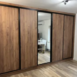 Duża szafa wnękowa z przesuwnymi drzwiami o drewnianej fakturze i czarnych obramowaniach, z lustrzanym panelem, odbijającym wnętrze pokoju z biurkiem i wiertarką na podłodze.