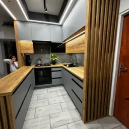 Nowoczesna kuchnia w odcieniach szarości i drewna z drewnianymi lamelami, oświetleniem LED podszafkowym i sufitowym, oraz blatem z naturalnego drewna. Na blacie czajnik, ekspres do kawy i roślina...