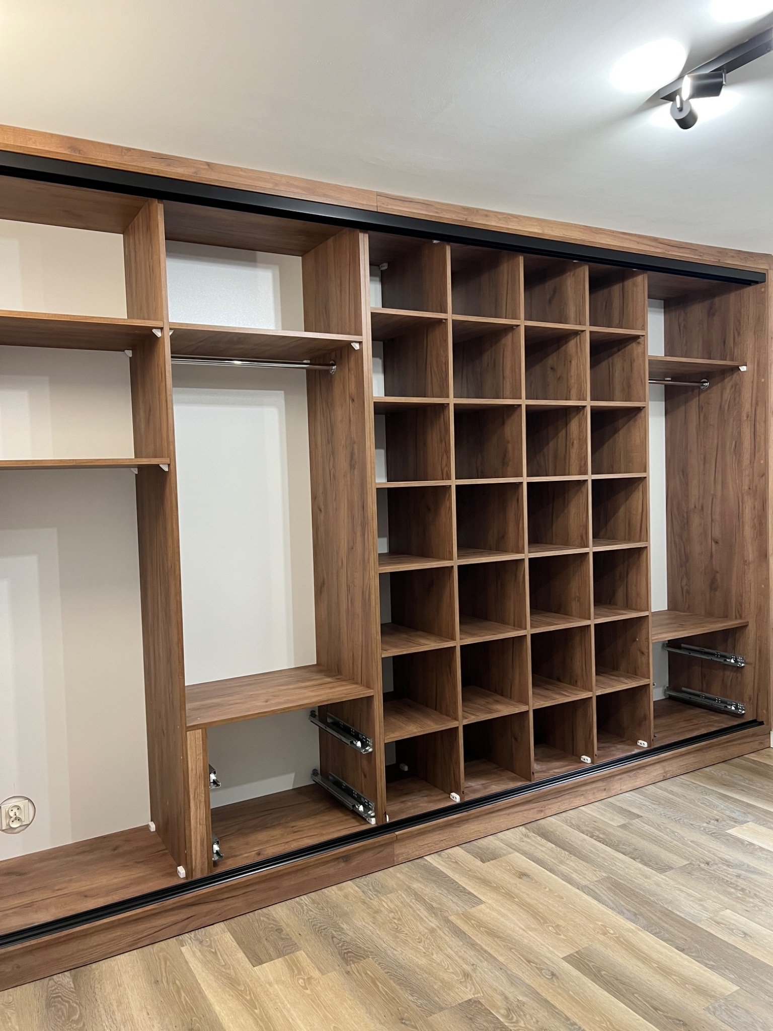 Wnętrze brązowej szafy przesuwnej z półkami i drążkami, widoczne prowadnice szuflad, na podłodze panele imitujące drewno.