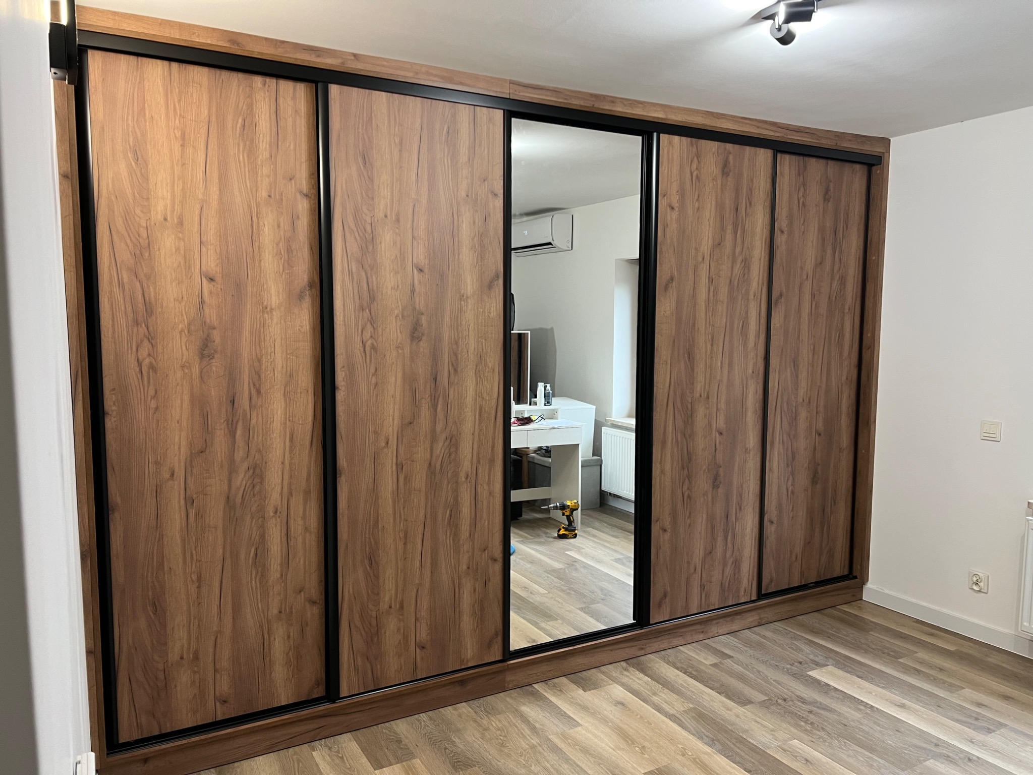 Duża szafa wnękowa z przesuwnymi drzwiami o drewnianej fakturze i czarnych obramowaniach, z lustrzanym panelem, odbijającym wnętrze pokoju z biurkiem i wiertarką na podłodze.