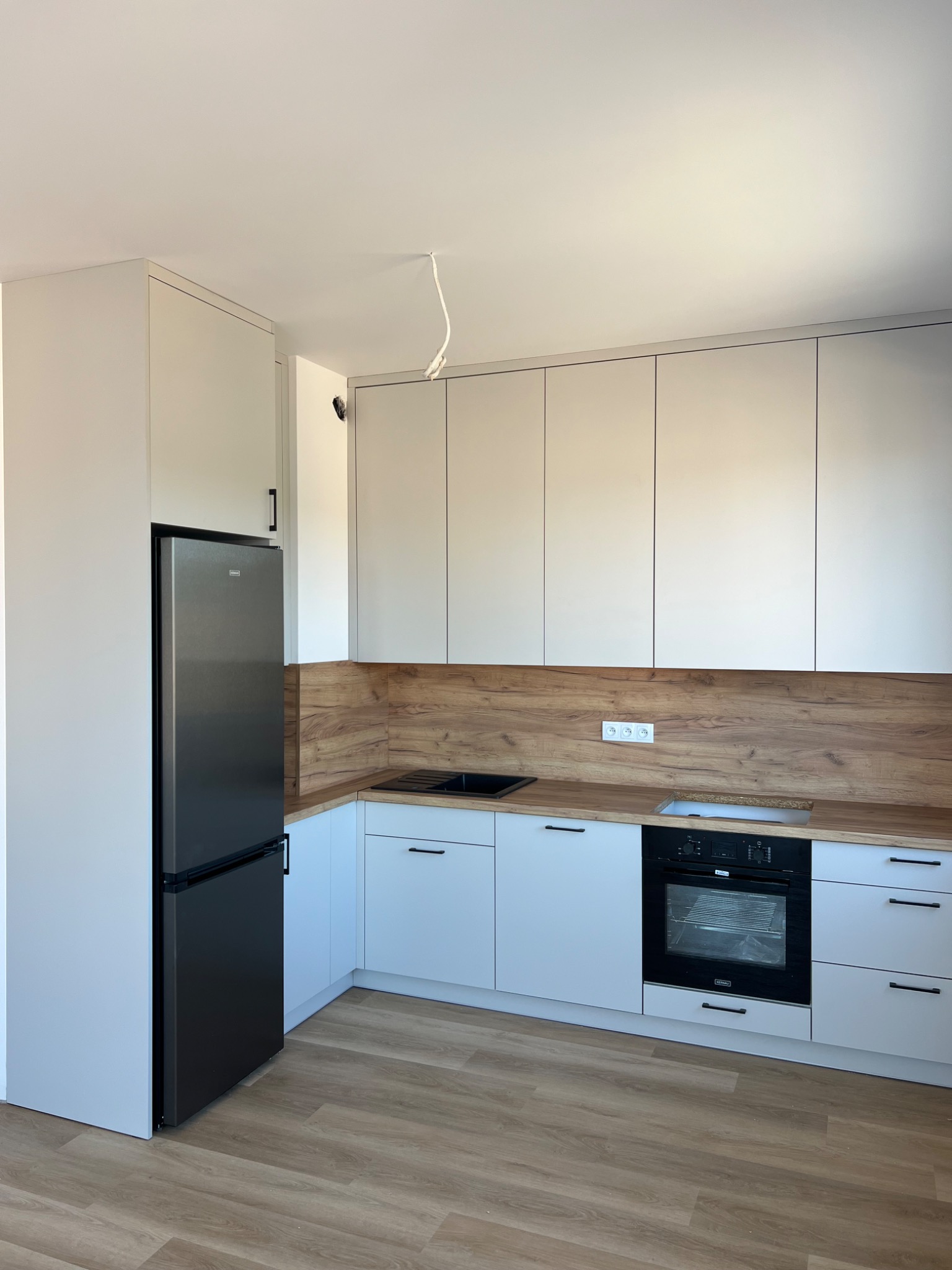 Jasna kuchnia z drewnianym blatem i panelem ściennym, czarny piekarnik i lodówka, minimalistyczne uchwyty, kabel zwisający z sufitu.