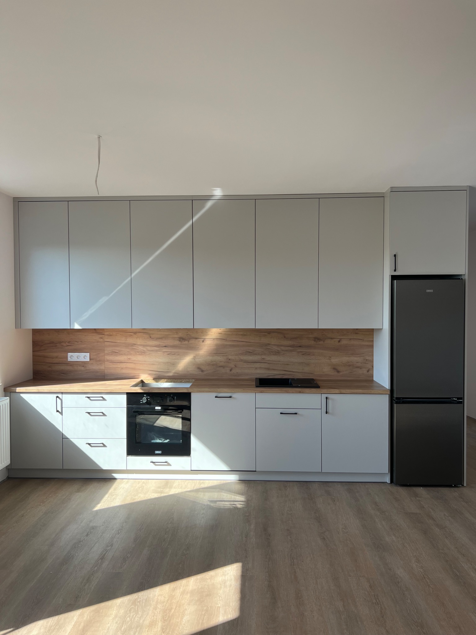 Nowoczesna kuchnia z szarymi szafkami, drewnianym blatem i panelem, czarnym piekarnikiem i lodówką, oświetlona promieniami słońca padającymi na podłogę.
