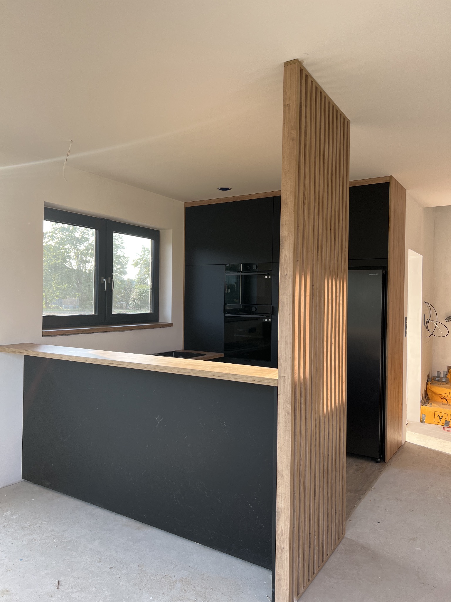 Częściowo ukończona, nowoczesna kuchnia z czarnymi szafkami, drewnianym blatem i ażurową, drewnianą ścianką działową, widoczna w jasnym wnętrzu w trakcie wykończenia.