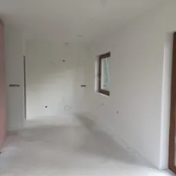 Wnętrze w trakcie wykańczania: świeżo wyszpachlowane ściany, surowy beton na podłodze, przygotowane kable elektryczne, obudowa kominka z izolacją, okna z widokiem na drzewa.