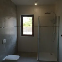 Nowoczesna łazienka z szarymi płytkami, kabiną prysznicową typu walk-in i podwieszaną toaletą, okno z widokiem na zieleń.