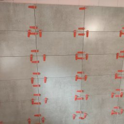 Remontowo budowlana - Układanie płytek w łazience z użyciem czerwonych klipsów poziomujących, widoczny fragment ściany z szarymi płytkami i gniazdko elektryczne.