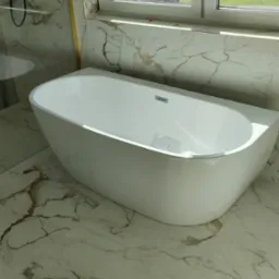 Nowoczesna, wolnostojąca wanna z minimalistyczną armaturą, umieszczona w łazience wykończonej marmurowymi płytkami, z widokiem na zielone pola za oknem.