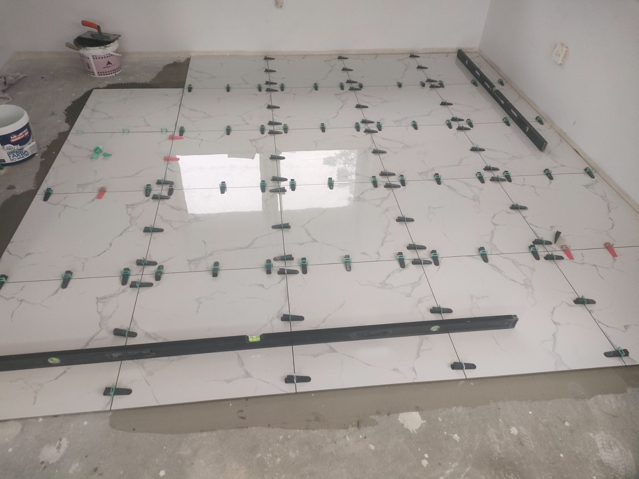 Układanie białych płytek podłogowych z marmurowym wzorem, poziomnica na płytkach, system poziomowania płytek z czarnymi i czerwonymi klinami, wiadro z klejem w tle.