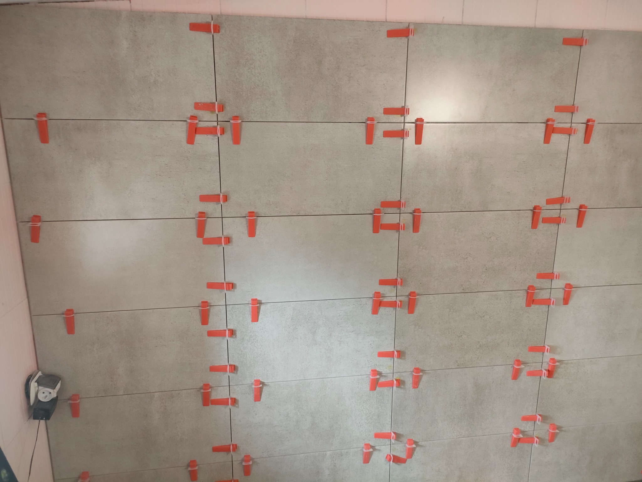 Układanie płytek w łazience z użyciem czerwonych klipsów poziomujących, widoczny fragment ściany z szarymi płytkami i gniazdko elektryczne.