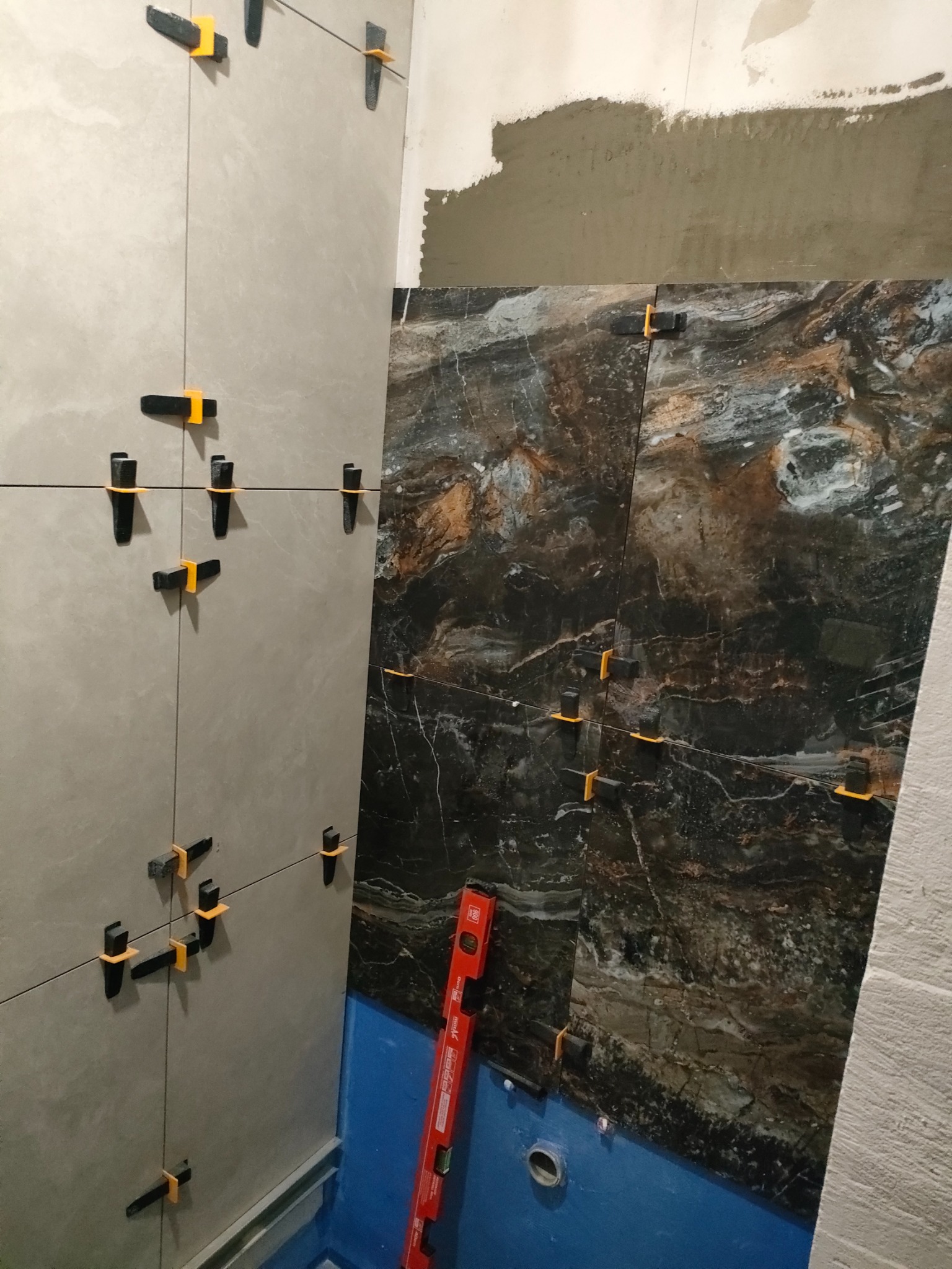 Montaż płytek w łazience: płytki imitujące marmur na ścianie obok jasnych płytek ceramicznych, użycie klipsów poziomujących, widoczna poziomica i fragment hydroizolacji.