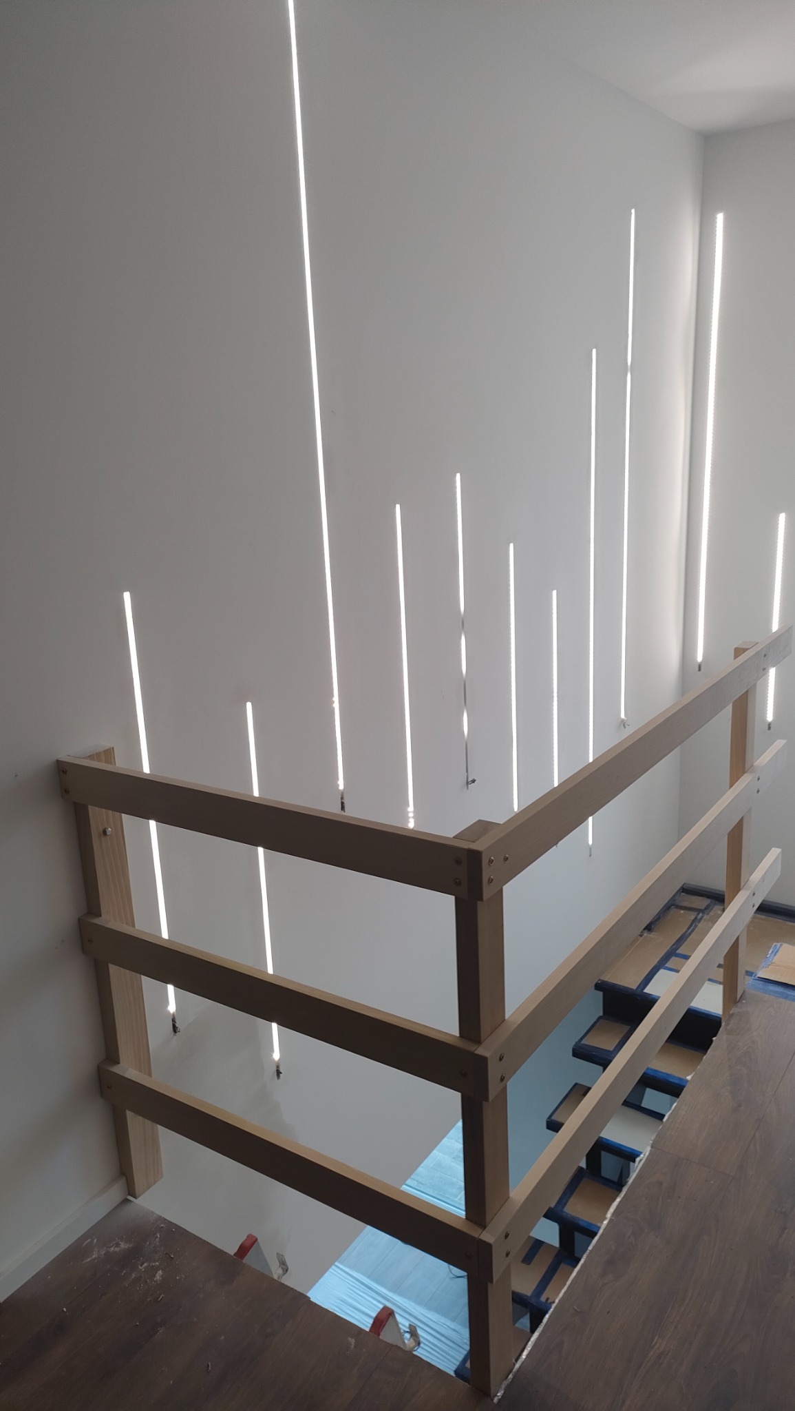 Wnętrze w trakcie prac wykończeniowych: drewniana balustrada schodów na tle białej ściany z pionowymi lampami LED, widoczne zabezpieczone schody.