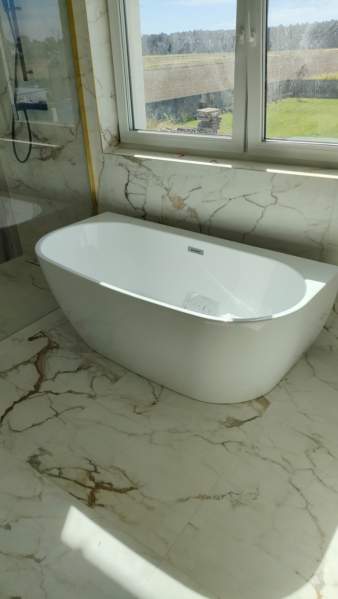 Nowoczesna, wolnostojąca wanna z minimalistyczną armaturą, umieszczona w łazience wykończonej marmurowymi płytkami, z widokiem na zielone pola za oknem.
