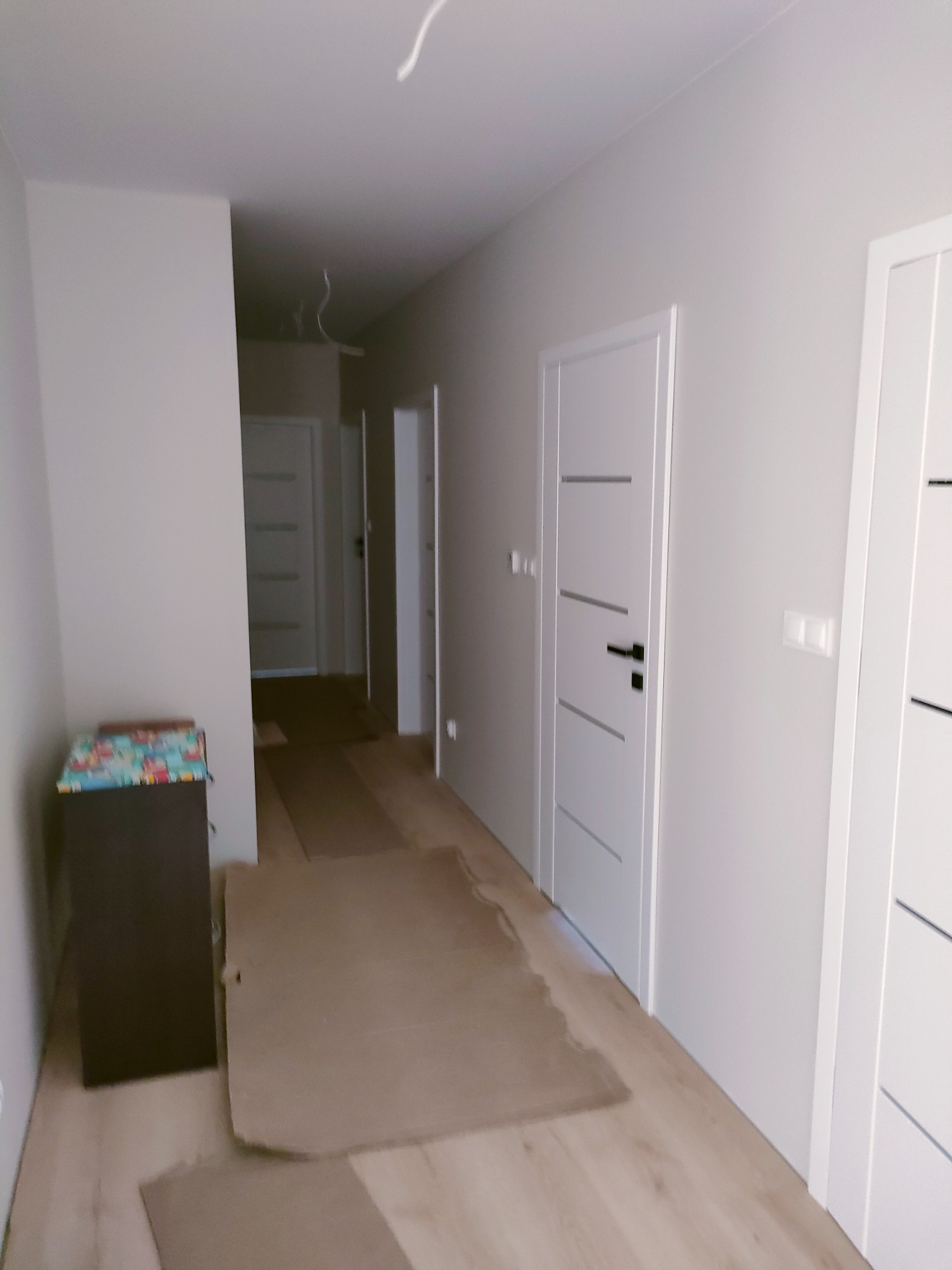 Korytarz w trakcie prac wykończeniowych z zabezpieczoną podłogą kartonem, widoczne białe drzwi z poziomymi frezami i szare ściany.