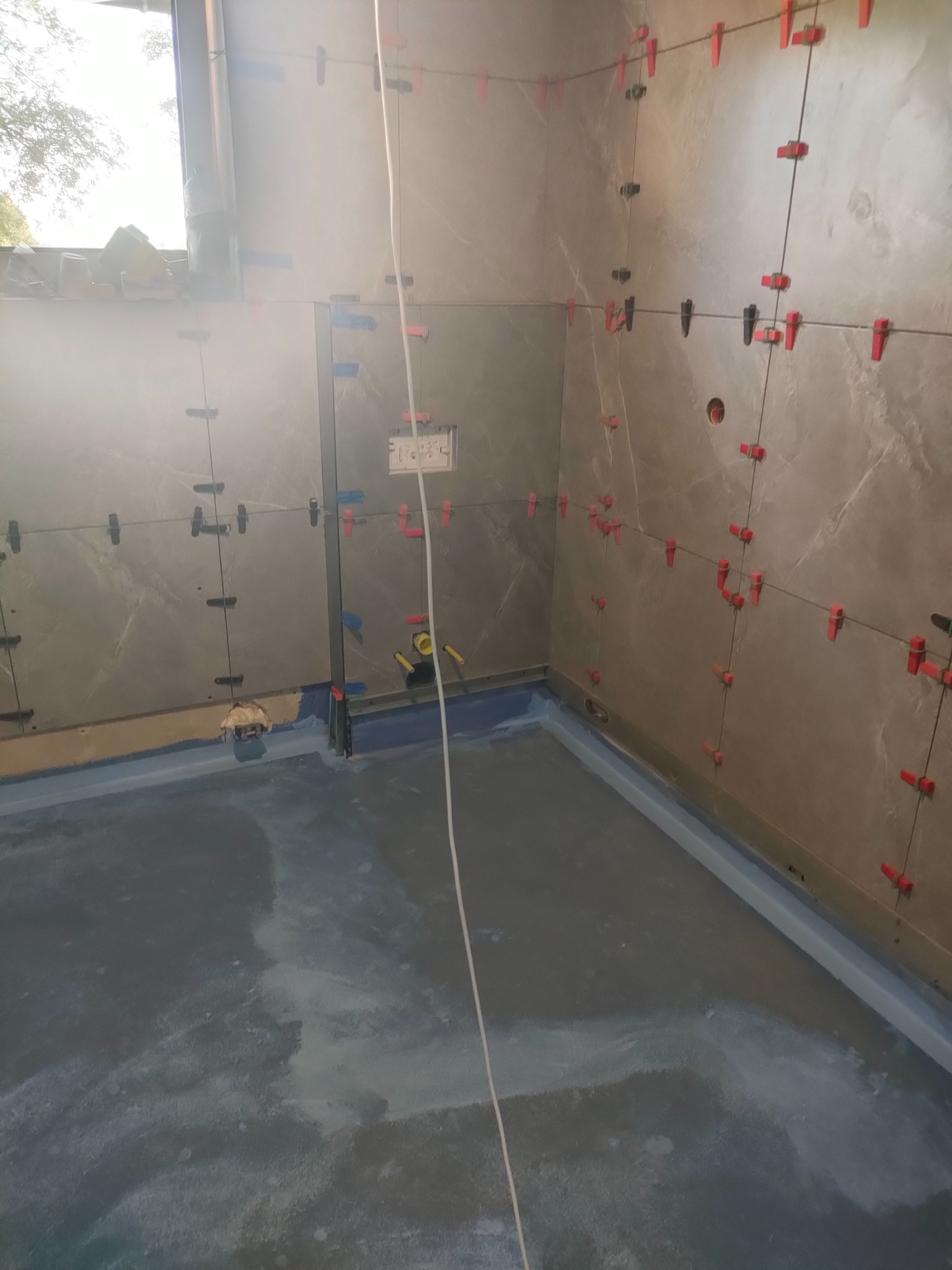 Łazienka w trakcie kładzenia płytek ściennych z użyciem systemu poziomowania; widoczne krzyżyki i kliny, przygotowane podłoże pod posadzkę.