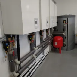 Instalacja z czterema białymi jednostkami wewnętrznymi, szary zbiornik akumulacyjny i czerwony zbiornik wyrównawczy, połączone rurami z izolacją, na tle szarej podłogi w pomieszczeniu technicznym.