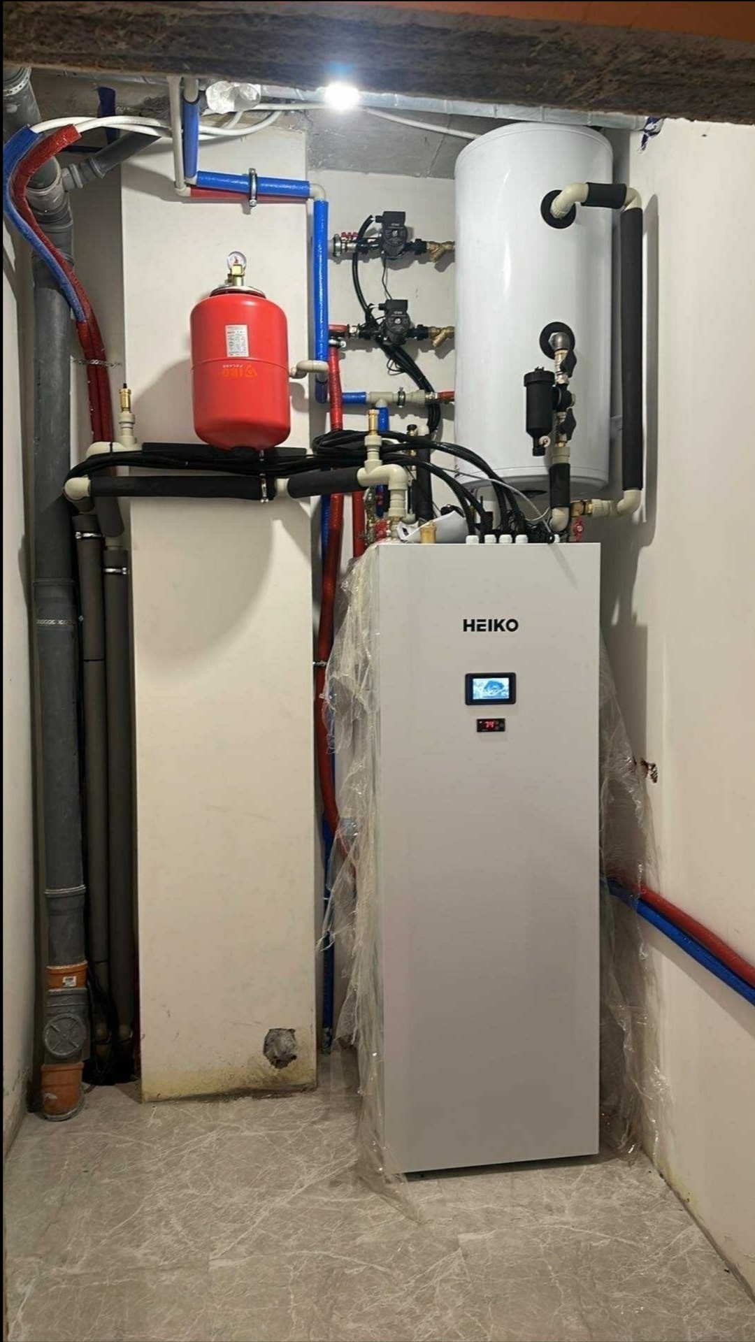 Zmontowana instalacja z pompą ciepła Heiko, zbiornikiem i orurowaniem w pomieszczeniu technicznym. Widoczne elementy instalacji i podłączenia.