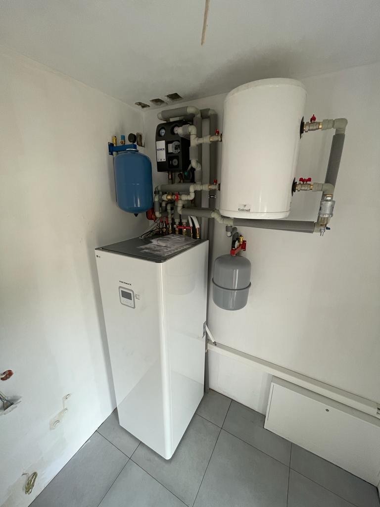 Biała pompa ciepła LG Therma V z zbiornikiem Galmet i instalacją hydrauliczną w kotłowni. Widoczne rury, zawory i osprzęt. Szare płytki podłogowe.