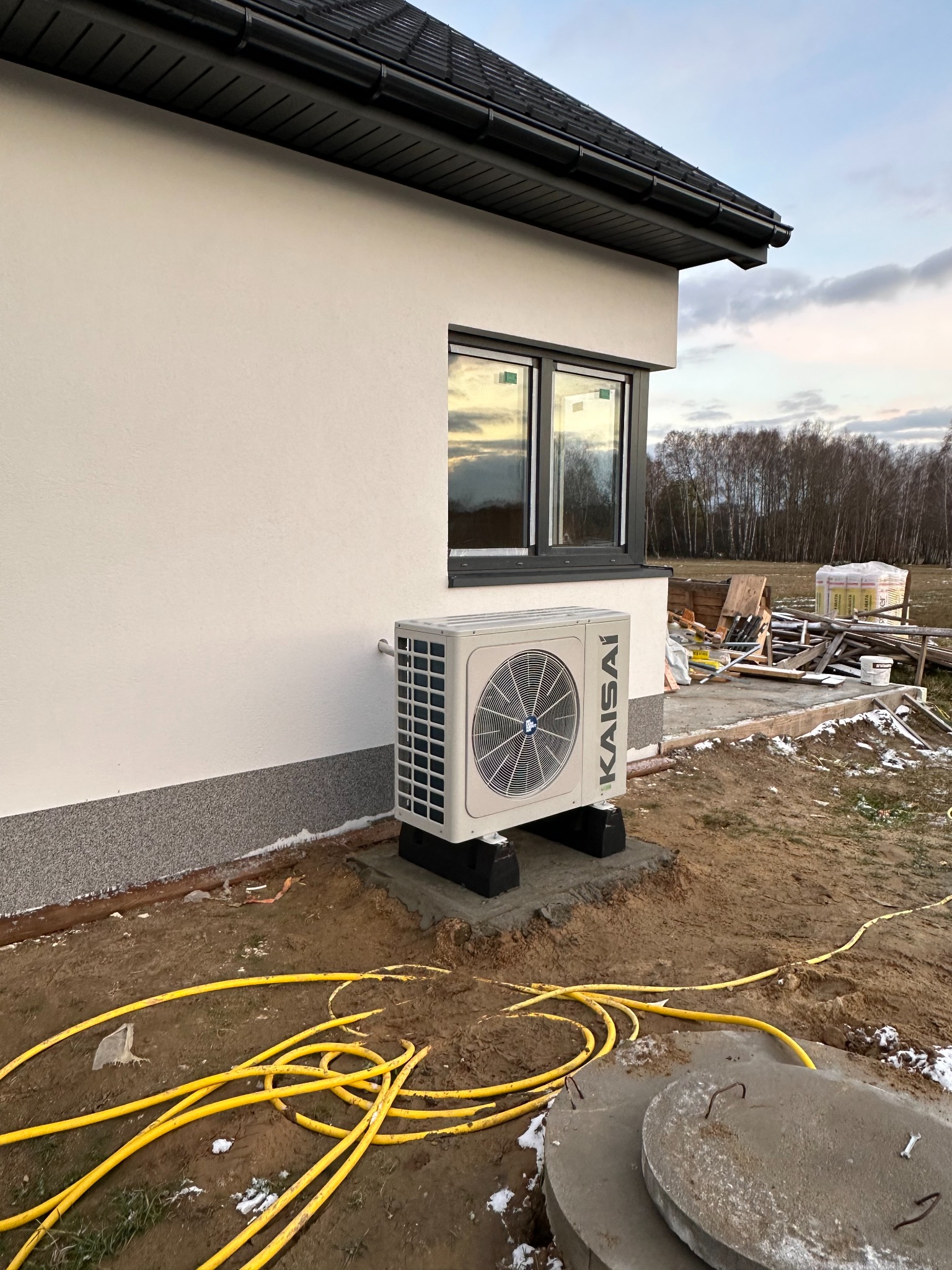 Biała pompa ciepła Kaisai zamontowana na betonowym fundamencie przy ścianie domu z szarą podmurówką. W tle okno z odbiciem nieba i lasu, oraz materiały budowlane.