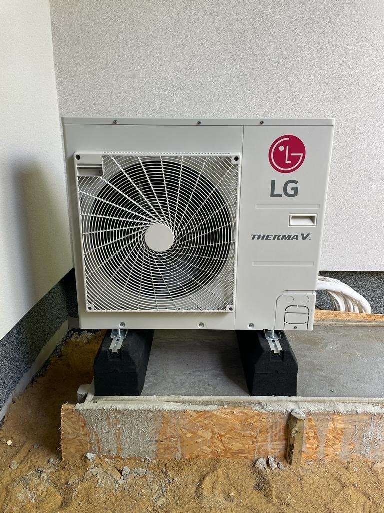 Biała zewnętrzna jednostka pompy ciepła LG Therma V, zamontowana na czarnych podkładkach, widoczna na tle surowego betonu i fragmentu drewnianej konstrukcji.