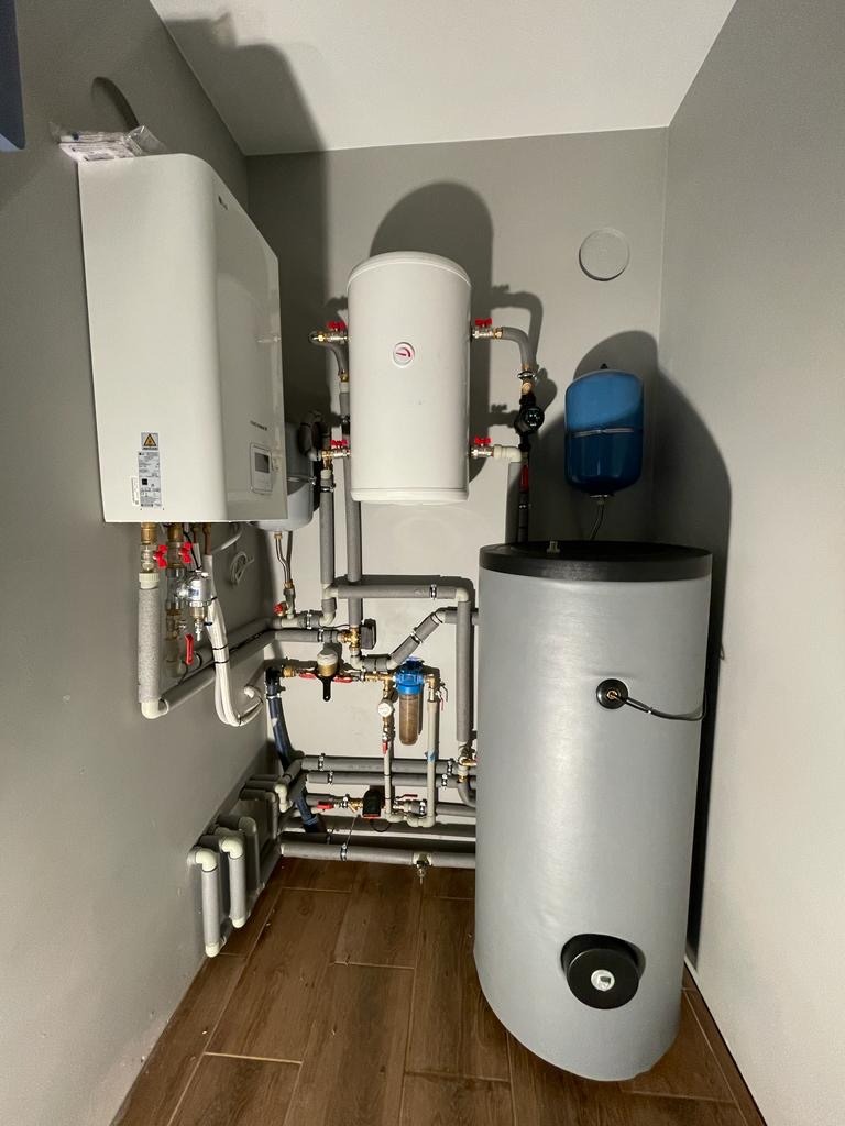 Zestaw urządzeń grzewczych w kotłowni: biały piec, zbiornik buforowy i szary zasobnik ciepłej wody, połączone rurami. Widoczne liczne zawory i filtry.