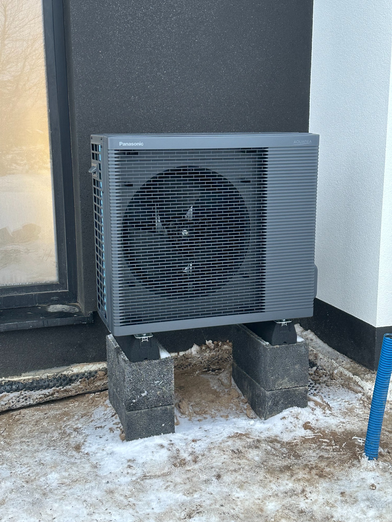 Szara pompa ciepła Panasonic Aquarea, zamontowana na betonowych bloczkach przy ścianie budynku w zimowej scenerii. Widoczny fragment okna i niebieska rura.