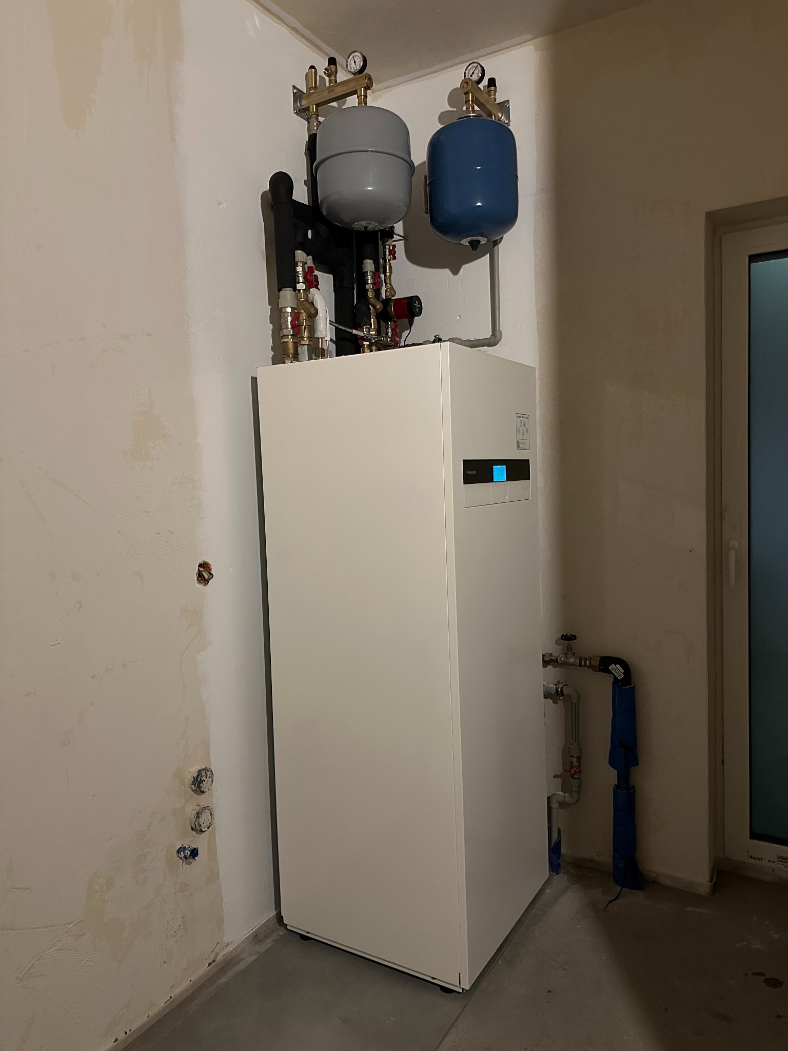 Biała pompa ciepła w surowym pomieszczeniu z szarym i niebieskim zbiornikiem wyrównawczym, widoczne rury i zawory. Ściany w trakcie wykończenia.