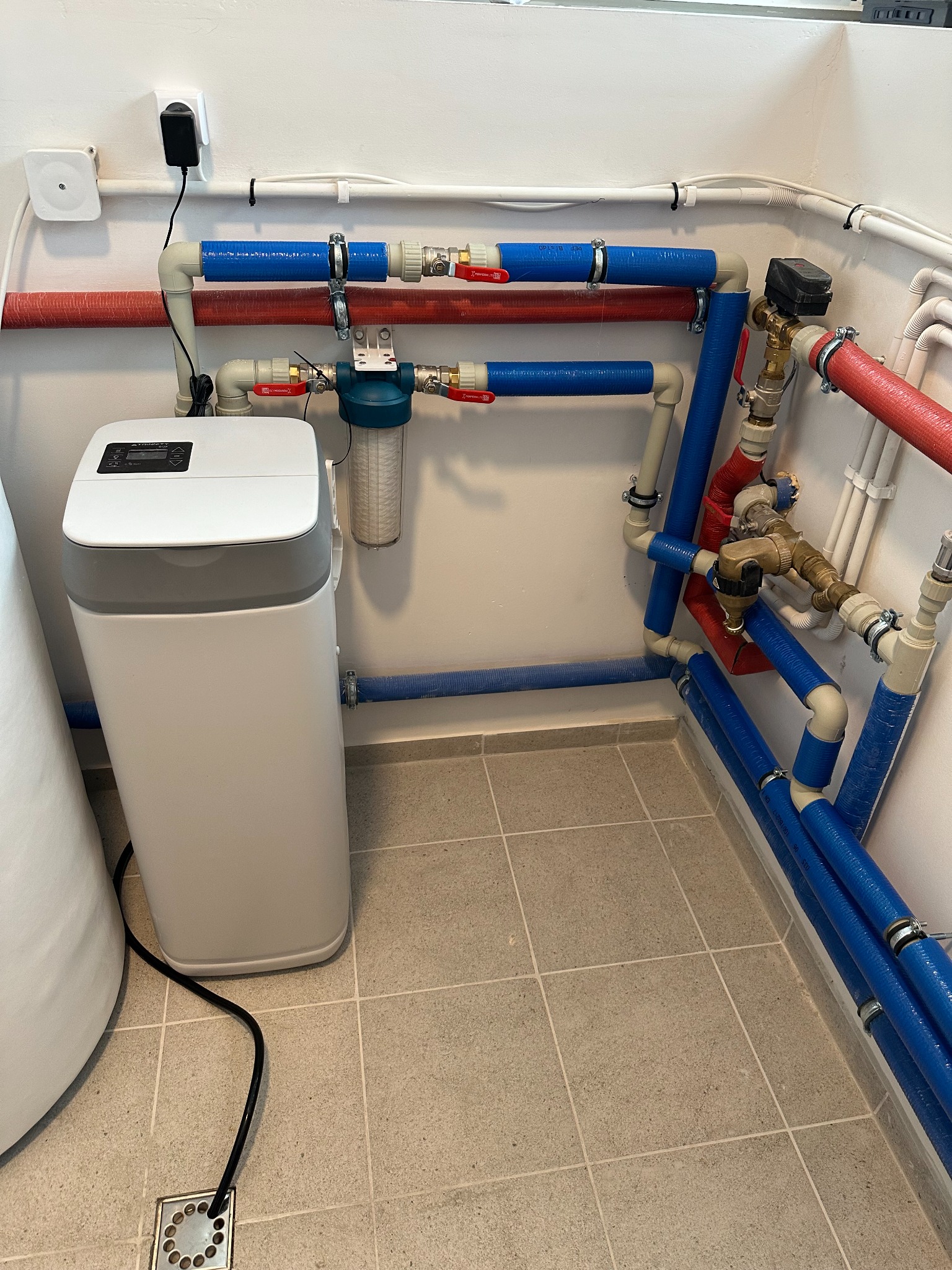 Nowoczesny system uzdatniania wody w pomieszczeniu technicznym. Biały filtr centralny z wyświetlaczem, niebieskie i czerwone rury z zaworami, szara płytka podłogowa.