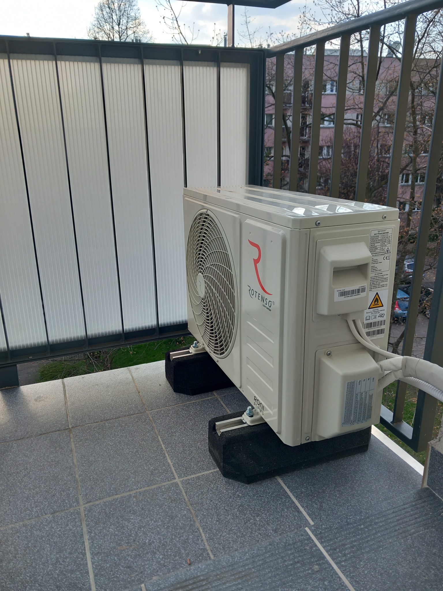 Klimatyzator Rotenso zamontowany na balkonie z szarą podłogą i metalową balustradą. Widok na budynek mieszkalny w tle. Urządzenie stoi na czarnych, gumowych podkładkach.