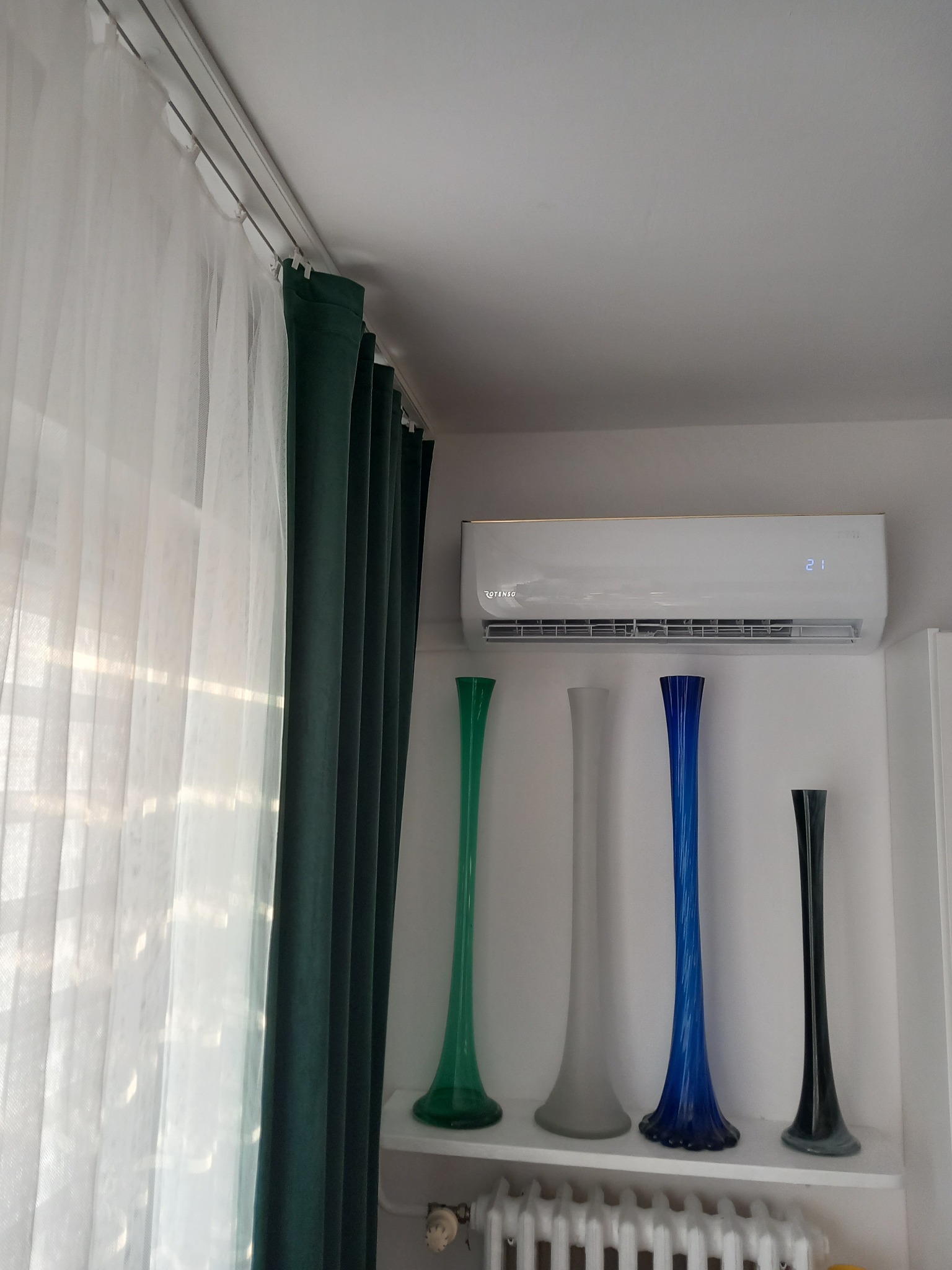 Biała klimatyzacja Rotenso na ścianie nad grzejnikiem i kolekcją wazonów. Obok okno z białymi firanami i zielonymi zasłonami. Ujęcie z dołu.