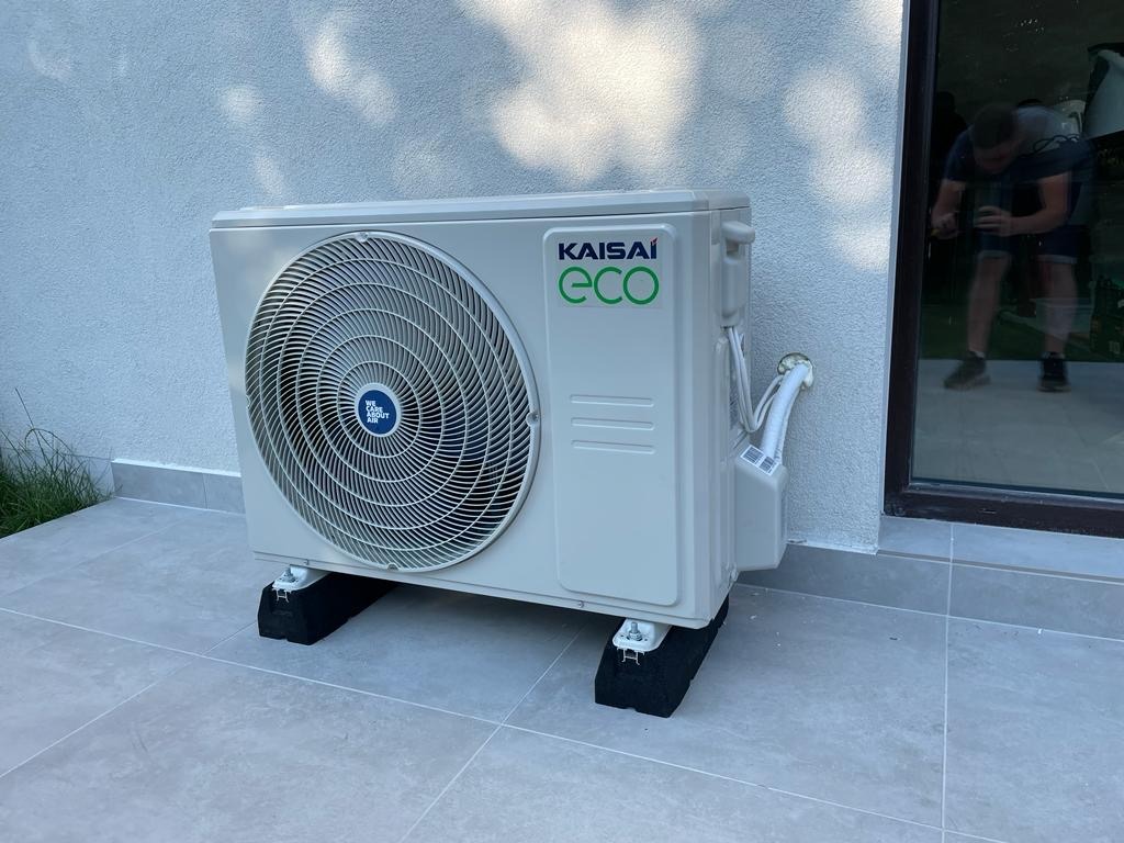 Biała jednostka zewnętrzna klimatyzacji Kaisai Eco, zamontowana na czarnych podkładkach, z widocznymi przewodami i fragmentem okna z odbiciem osoby.