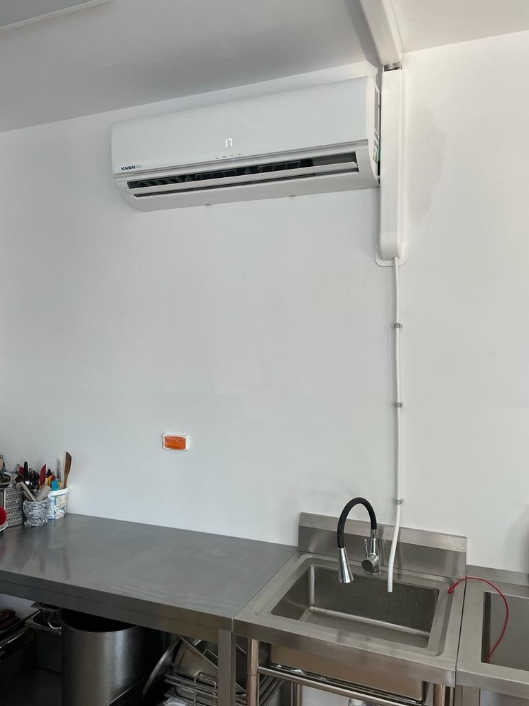 Biały klimatyzator Kaisai z wyświetlaczem temperatury (17°C) zamontowany na ścianie nad metalowym zlewem w pomieszczeniu o minimalistycznym wystroju.