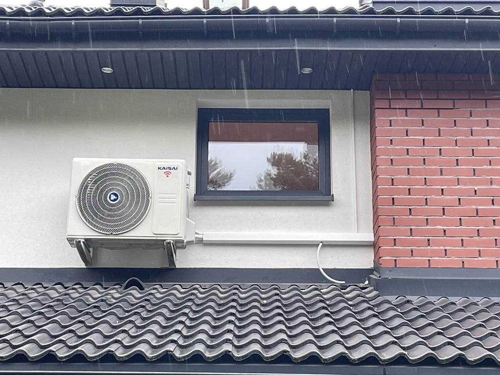 Biały klimatyzator Kaisai zamontowany na elewacji domu z ceglaną ścianą, widoczny z perspektywy dachu pokrytego ciemną dachówką. Deszczowa pogoda.
